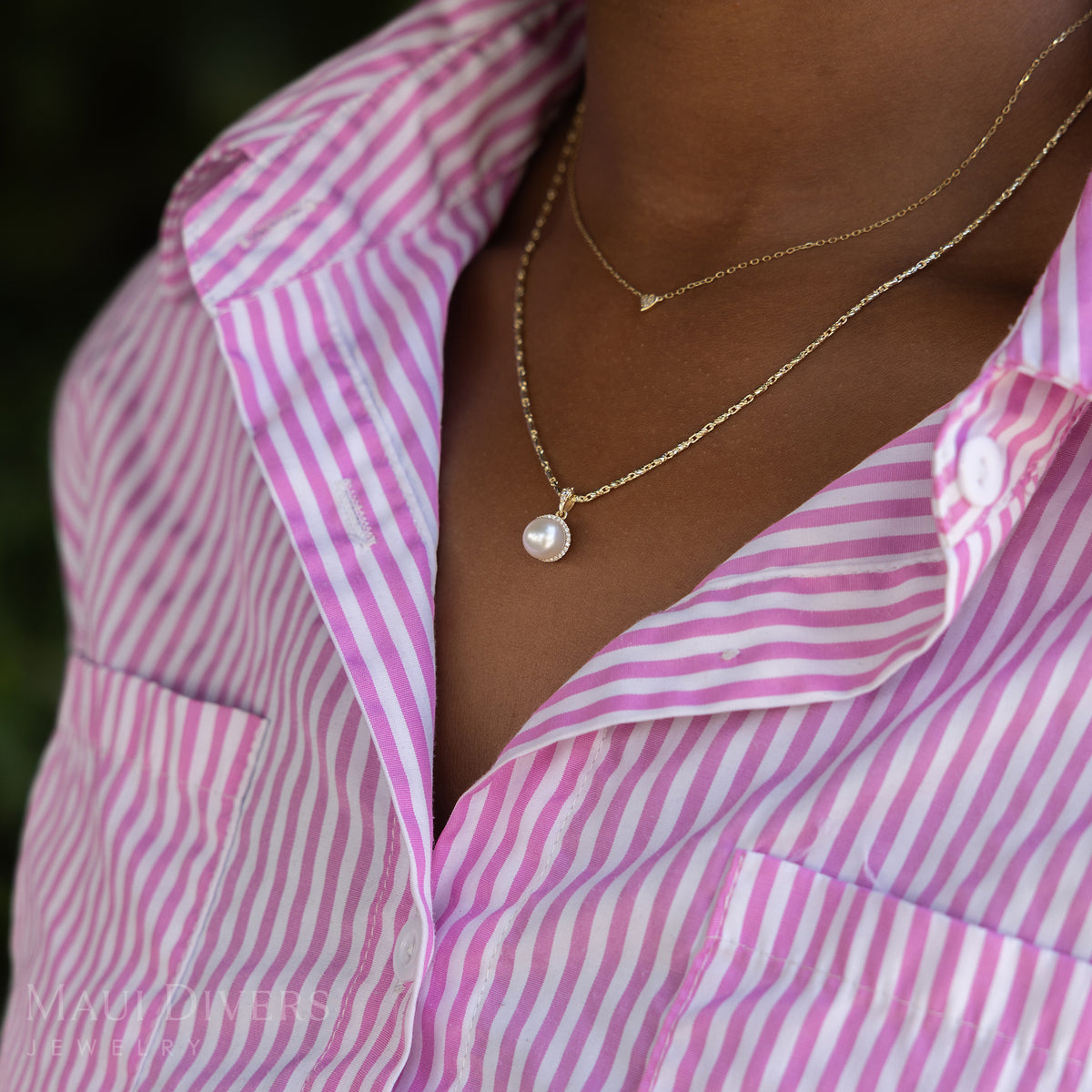 A side view close-up of a woman in a pink and white striped top wearing a Halo Akoya White Pearl Pendant in 14k yellow gold with diamonds on a raso chain, layered with a tiny diamond heart necklace, against a blurred leafy green background