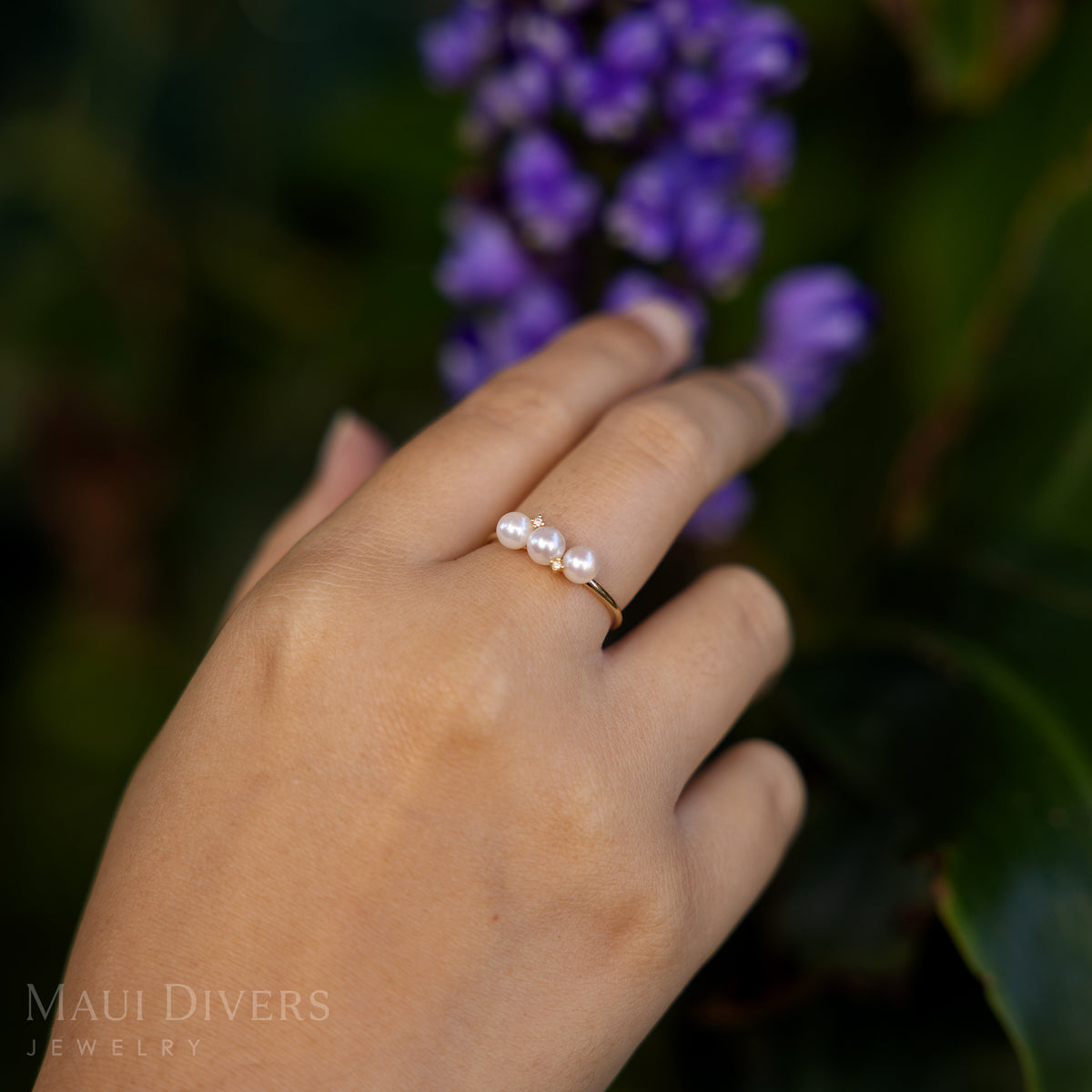 Close-up of a hand wearing an Akoya White Pearl Ring in 14k yellow gold with diamonds, against a blurred leafy green and purple flowers background