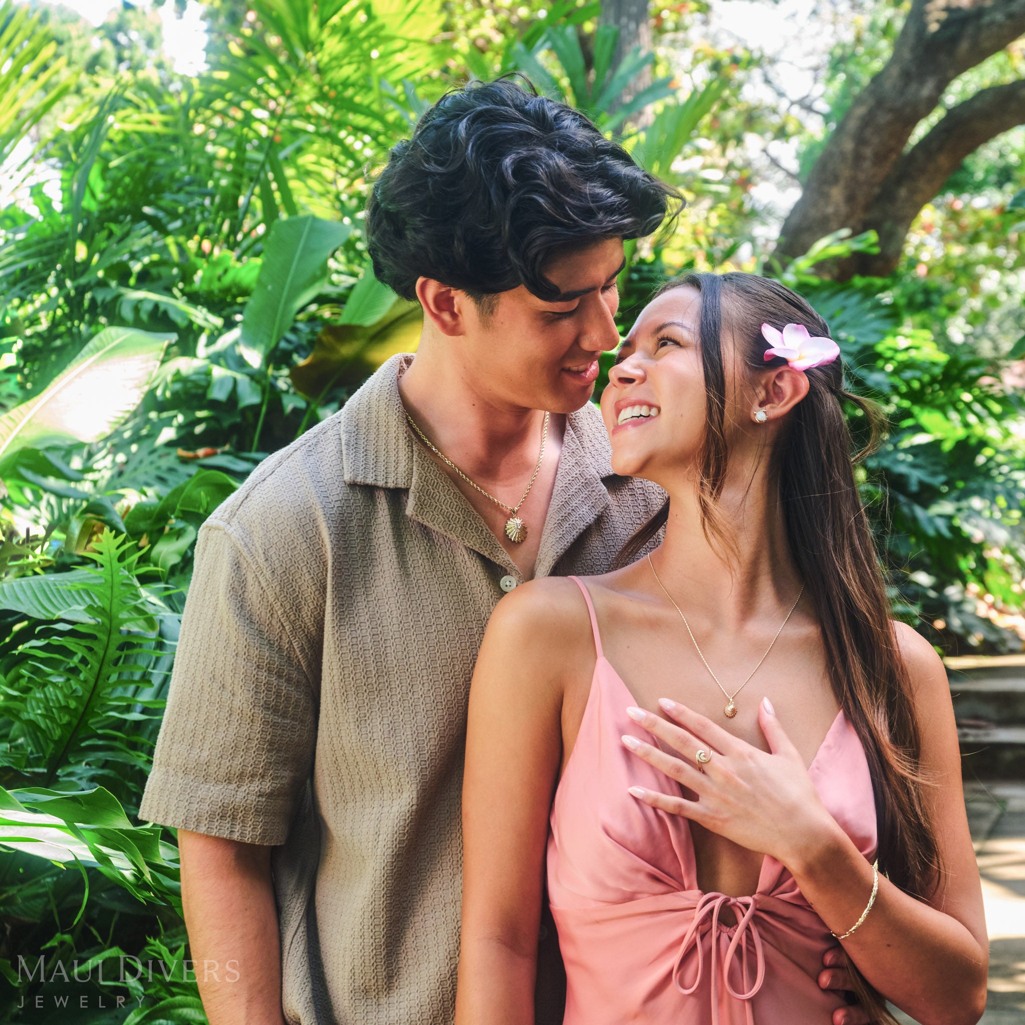 Smiling couple wearing matching ʻOpihi Pendants in 14k yellow gold with diamonds, with the woman also wearing a Nalu Ring in 14k yellow gold with diamonds, against a tropical forest background