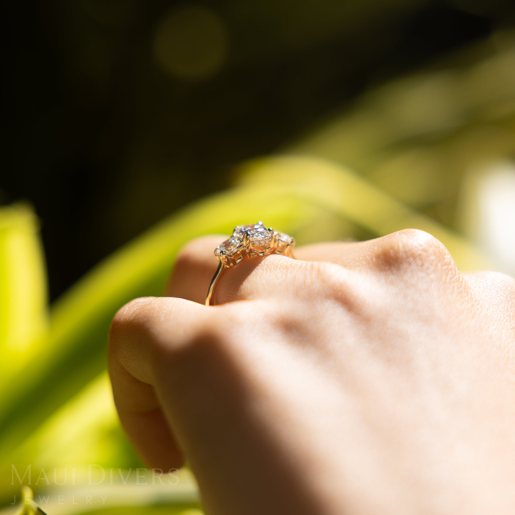 Hand wearing a gold ring with a lab-grown diamond, held against a blurred natural background