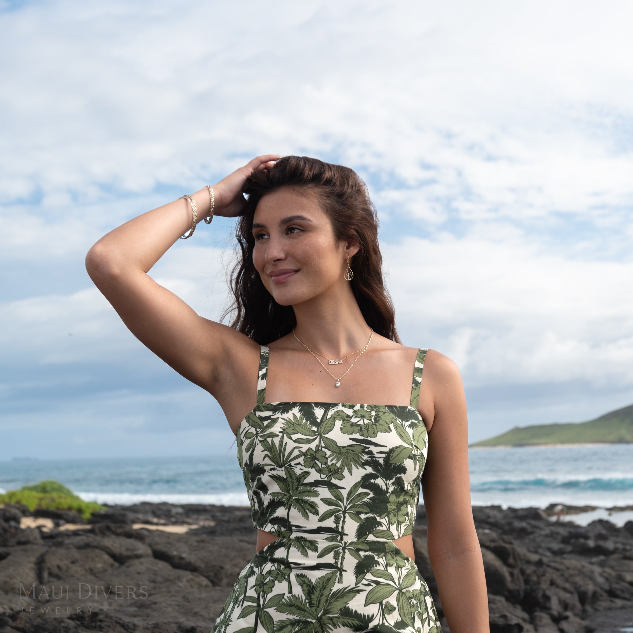 Woman in a green and white patterned dress wearing a lab-grown diamond pendant on a Singapore chain and a diamond Aloha necklace, standing on a rocky beach with ocean and sky in the background.