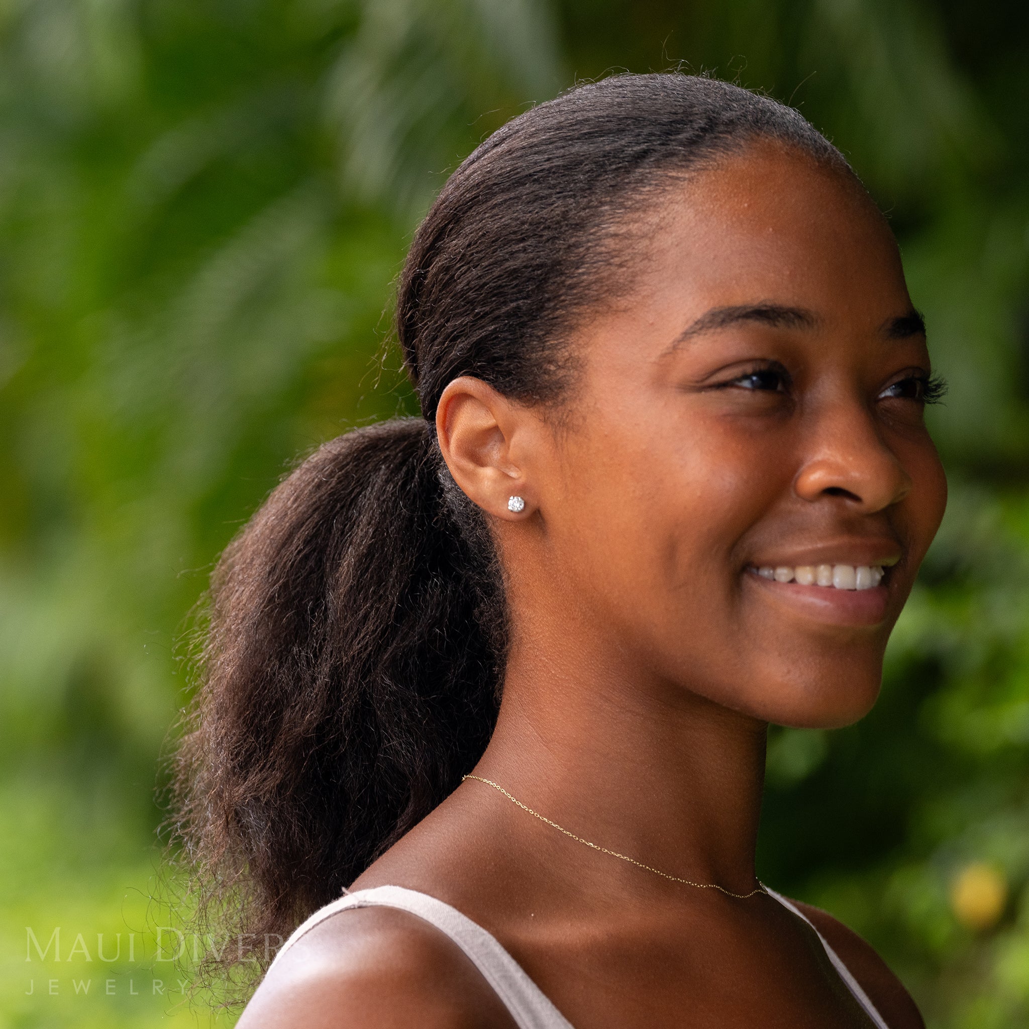 A smiling woman wearing a Hidden Hawaiʻi Maile Lab Grown Diamond Earrings in 14k White Gold on her ear, against a blurred leafy green background
