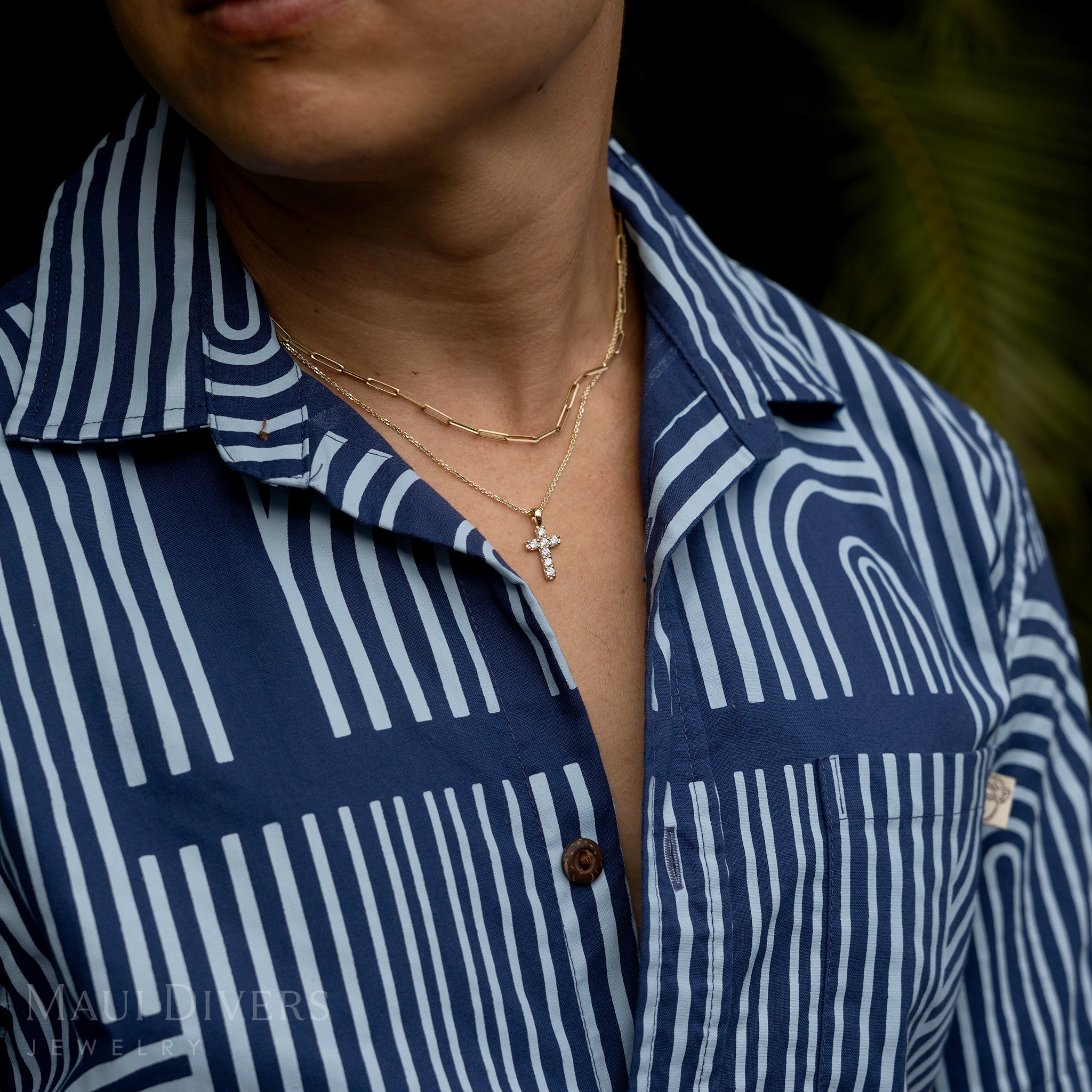A close-up of a guy in a two-toned blue patterned shirt wearing a Hidden Hawaiʻi Maile Lab Grown Diamond Cross Pendant in 14k yellow gold around his neck, layered with a paperclip chain against a blurred green leaf background