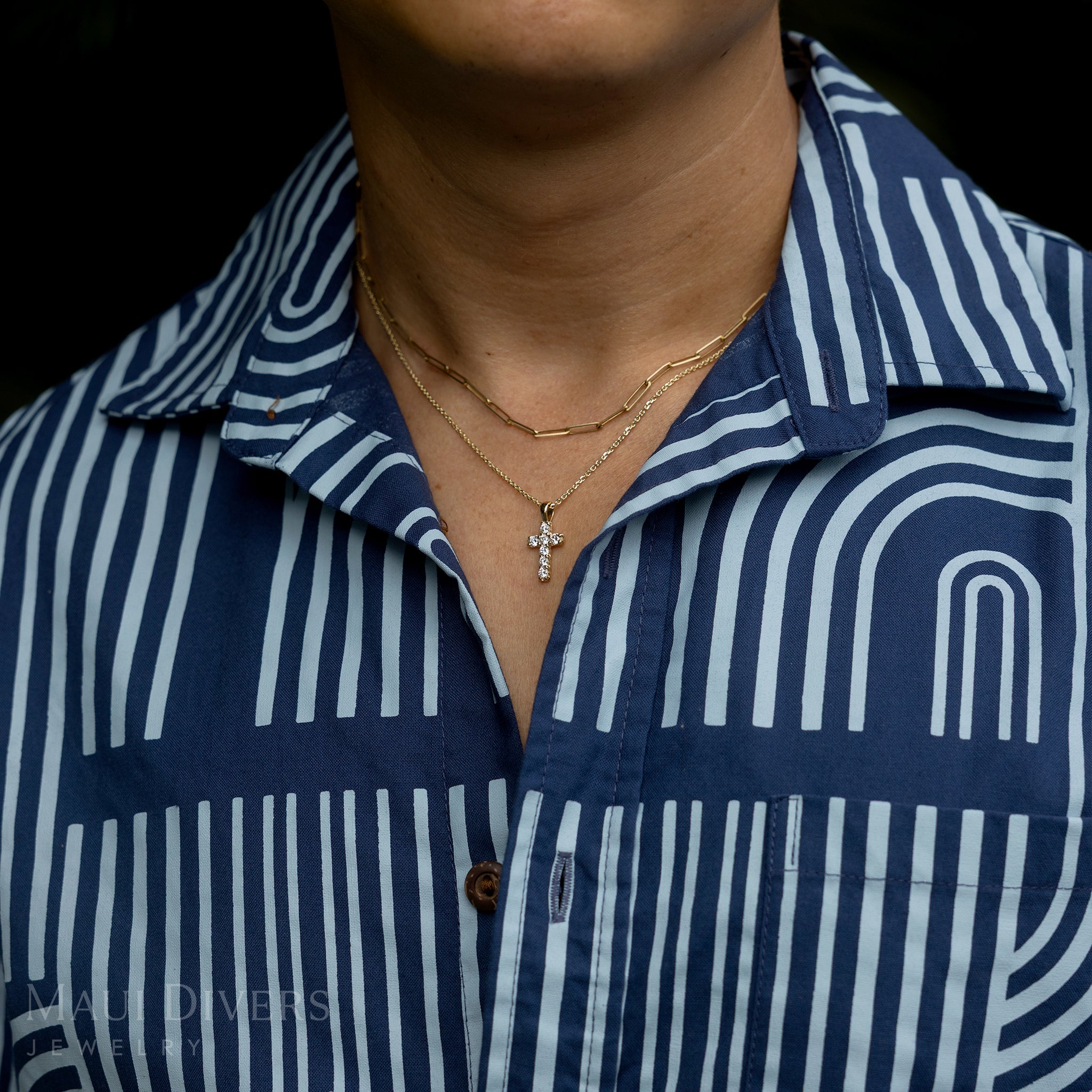 A close-up of a guy in a two-toned blue patterned shirt wearing a Hidden Hawaiʻi Maile Lab Grown Diamond Cross Pendant in 14k yellow gold around his neck, layered with a paperclip chain against a blurred green leaf background