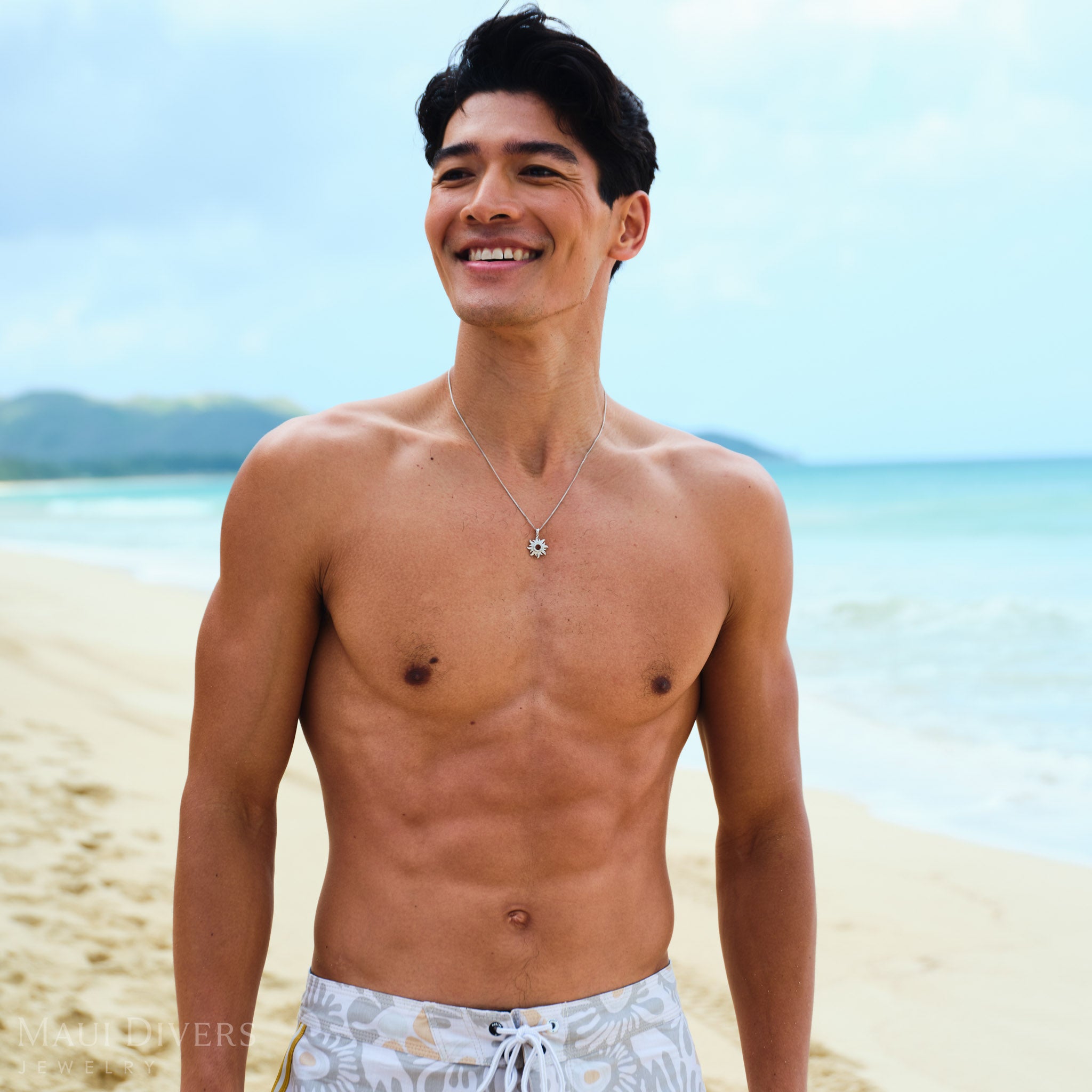 Man standing on a beach wearing a white gold sun pendant with a clear blue sky and ocean in the background