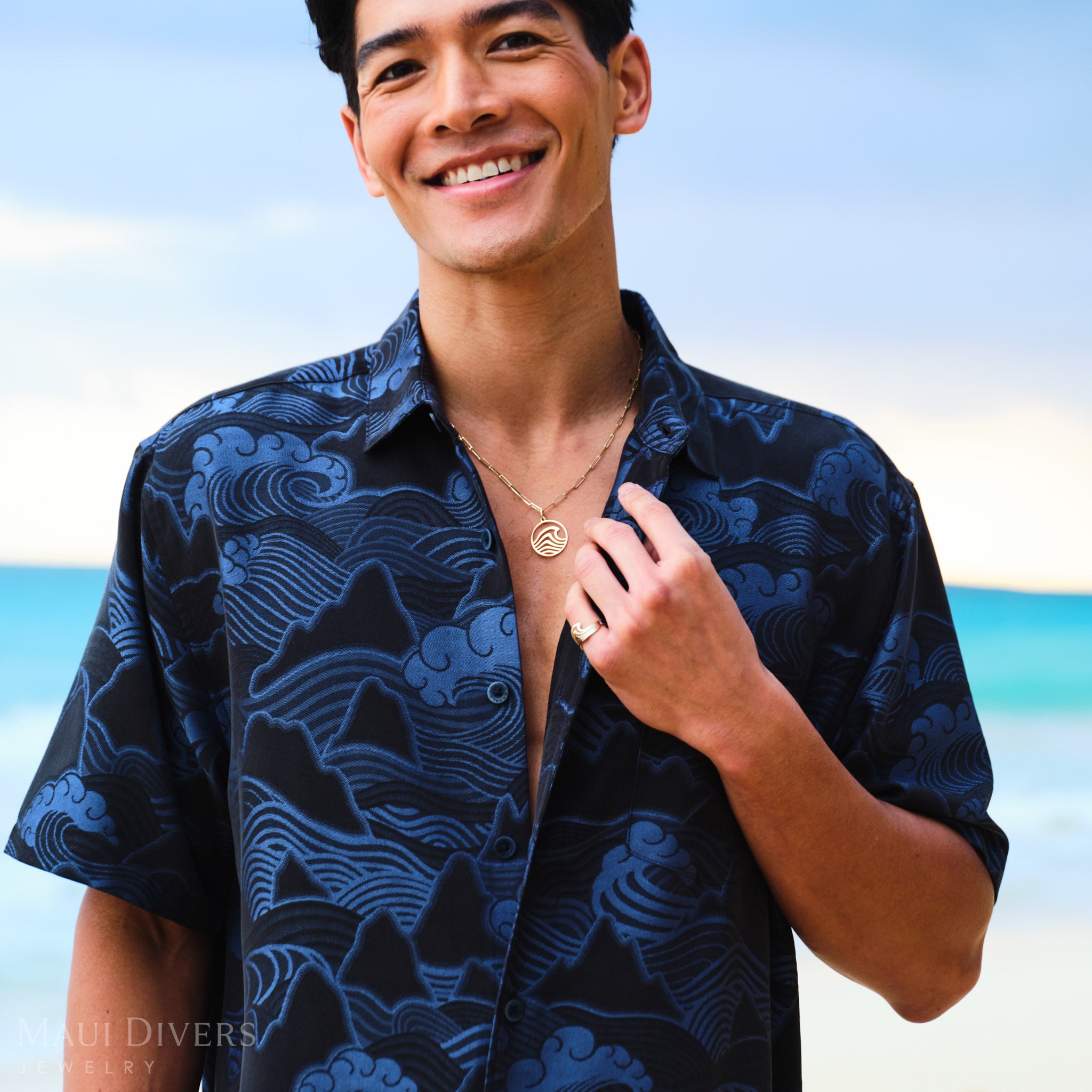 Man wearing a blue patterned shirt and a wave necklace and ring on a beach