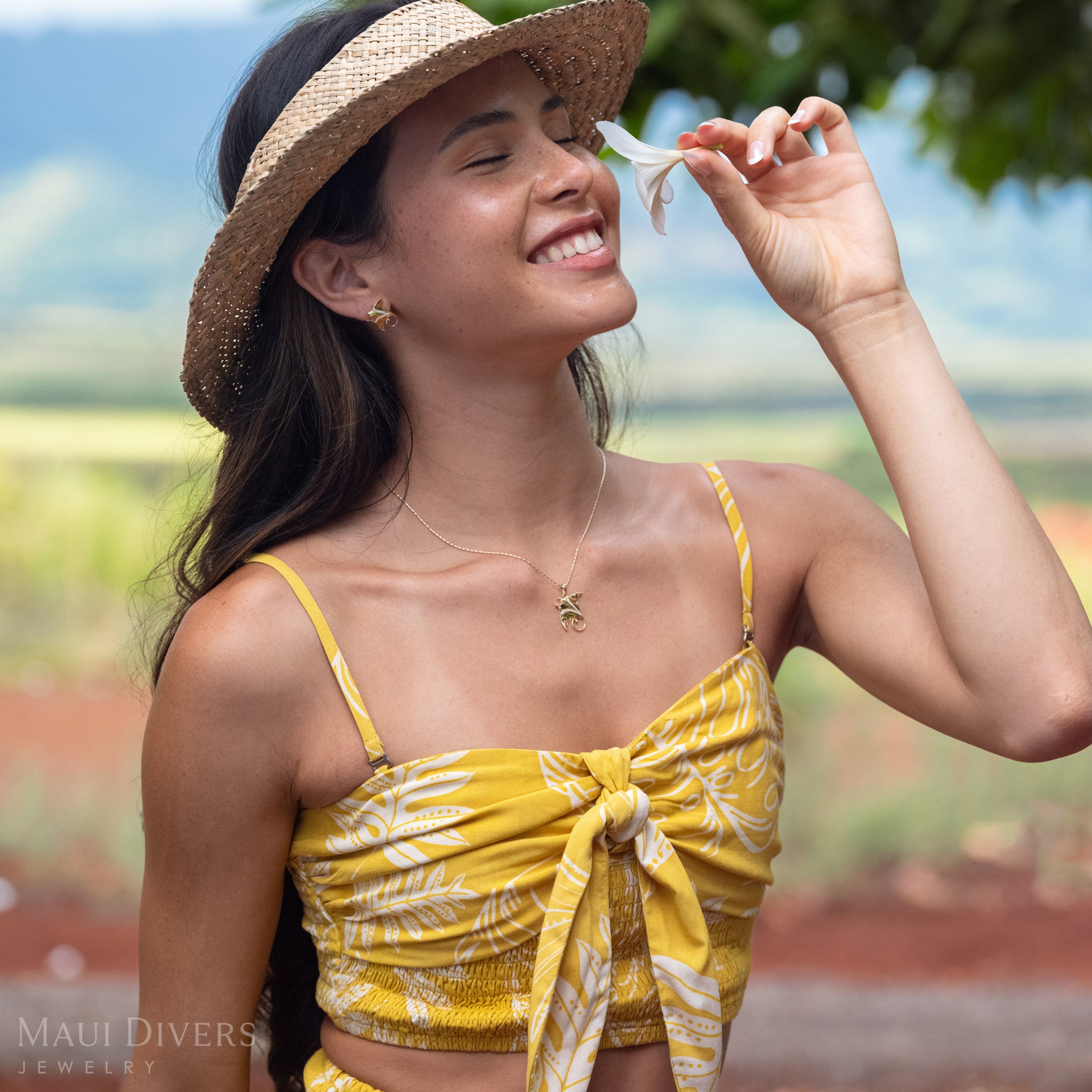 Woman smelling a white plumeria flower, wearing a 14k Gold Manta Ray Pendant around her neck with Natural Diamond detailing and matching earrings and wearing a brown woven hat. 