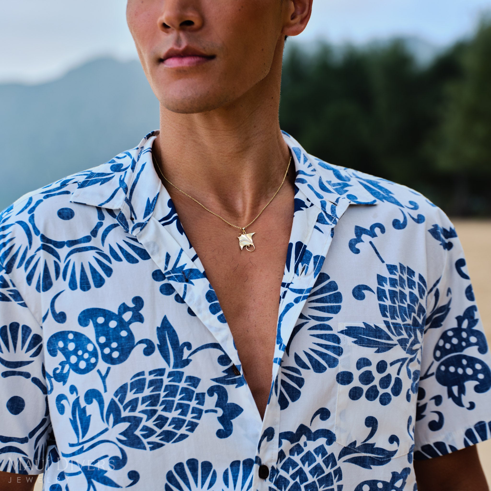 Man wearing a blue and white patterned shirt with a manta ray pendant on a rope chain outdoors.