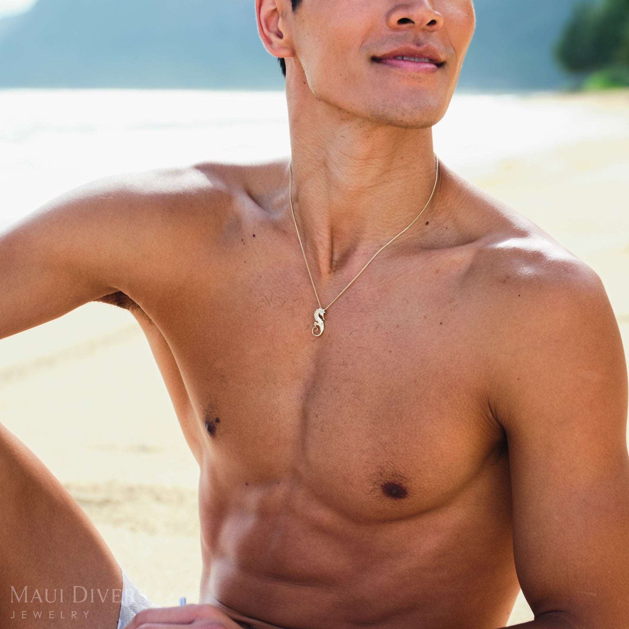 Man wearing a gold seahorse necklace on a beach.