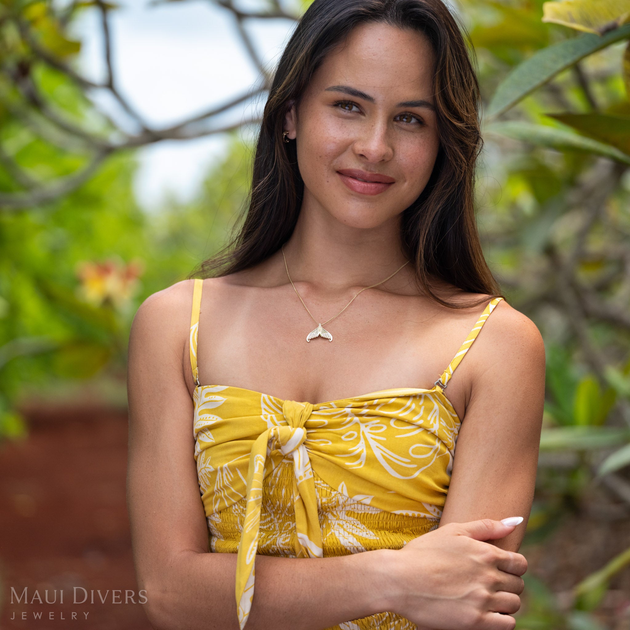 Woman with one arm crossed wearing a 14k Gold Mermaid Tail Necklace with Natural Diamond accents in front of a green natural background. 