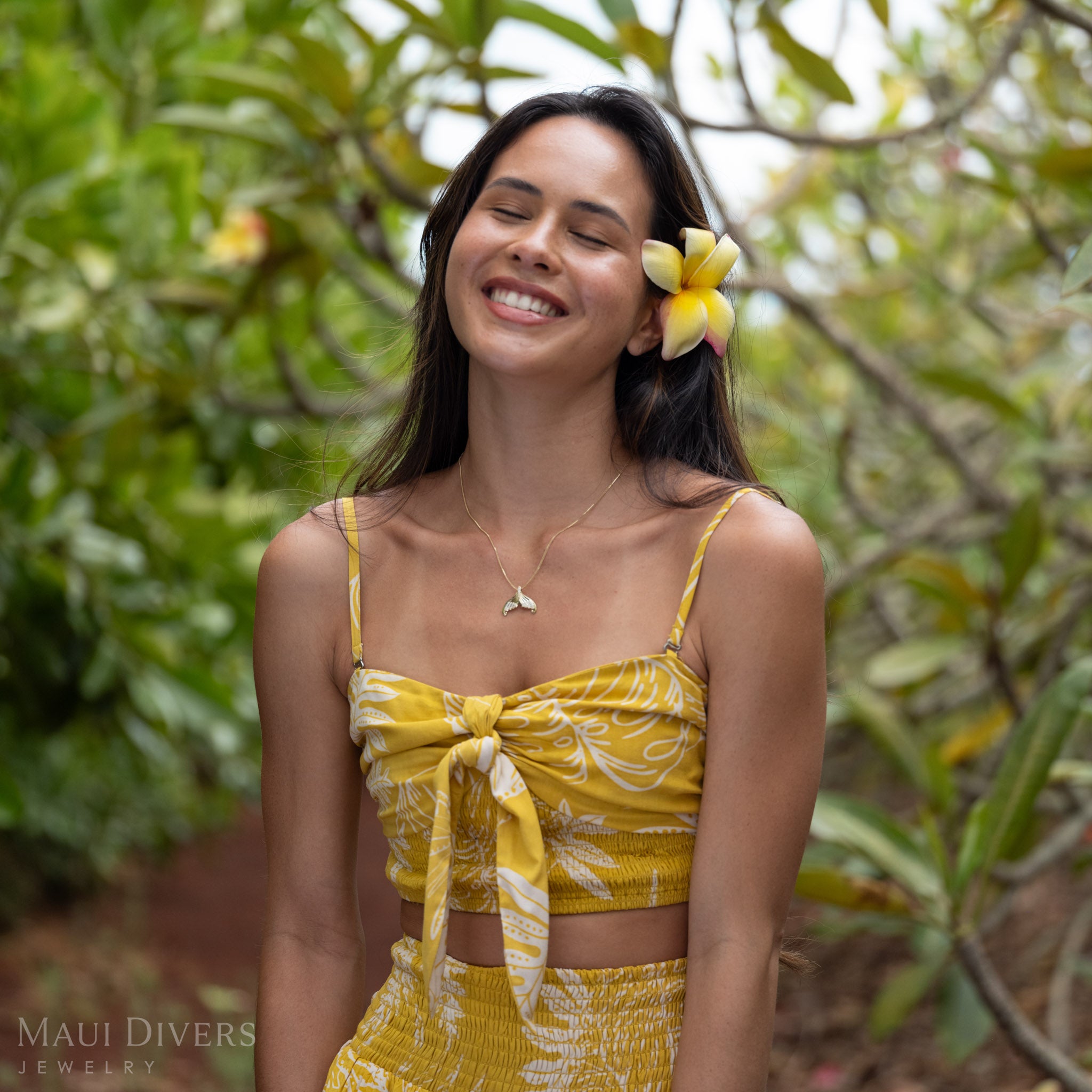 A woman smiling wearing a 14k Gold Mermaid Tail Necklace with Natural Diamond accents and a yellow flower in her hair in front of a green natural background. 