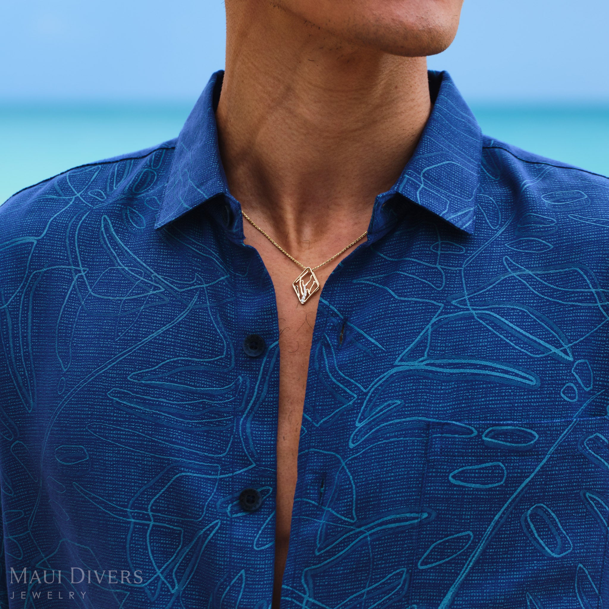 Man wearing a blue patterned shirt with a Koʻolau Mountain necklace against a blurred ocean background