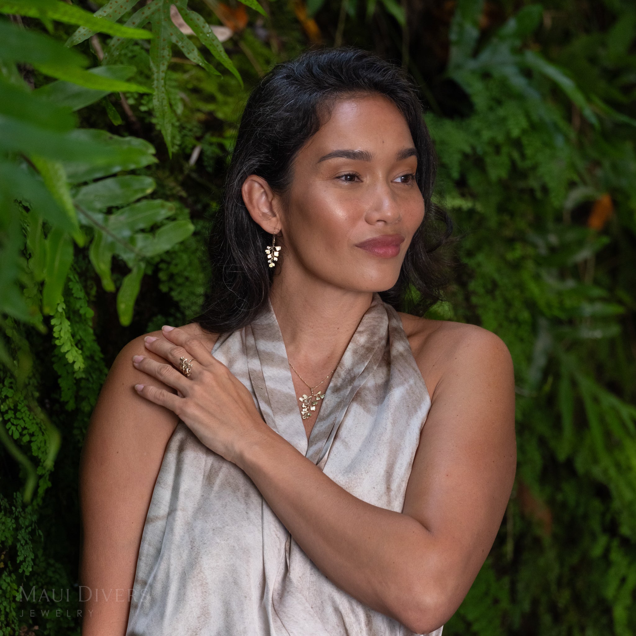 A woman with dark hair wearing a maiden hair fern pendant on a Singapore chain, a matching earring, a matching ring, a light beige and gray patterned halter top, standing in front of a lush green fern wall. 