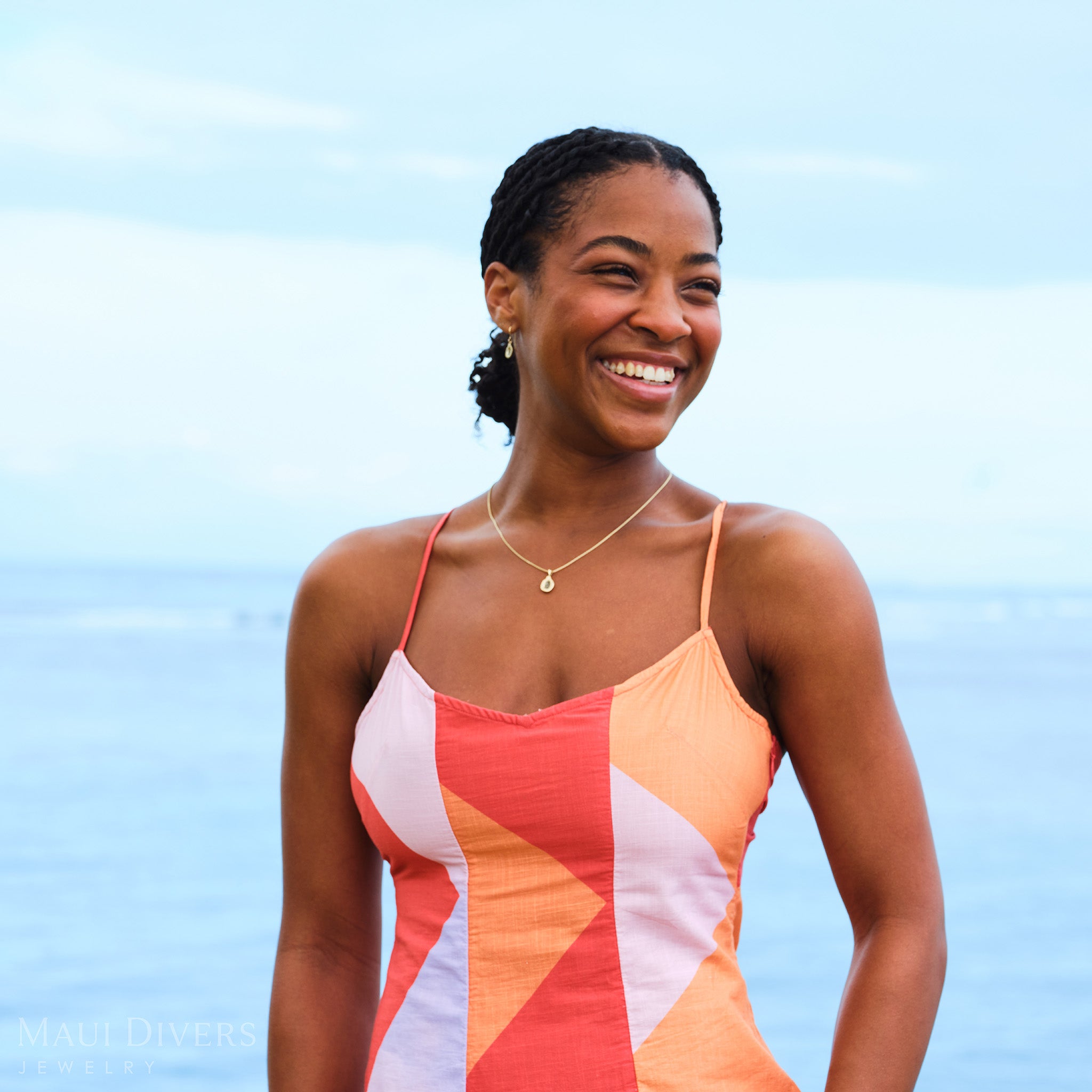 Smiling woman in an orange and pink top wearing a Cute Fruits Papaya Pendant in 14k yellow gold with black diamonds around her neck against a blurred ocean background