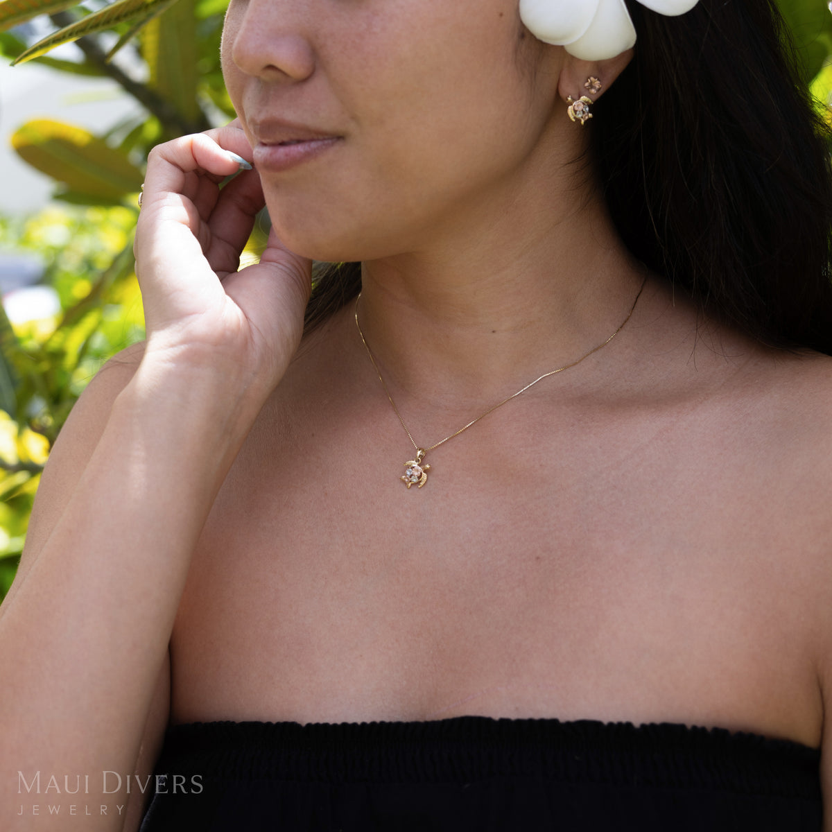 Close-up of a woman wearing a small gold sea turtle pendant with plumeria flowers in rose and white gold on a chain with matching earrings against a blurry natura background