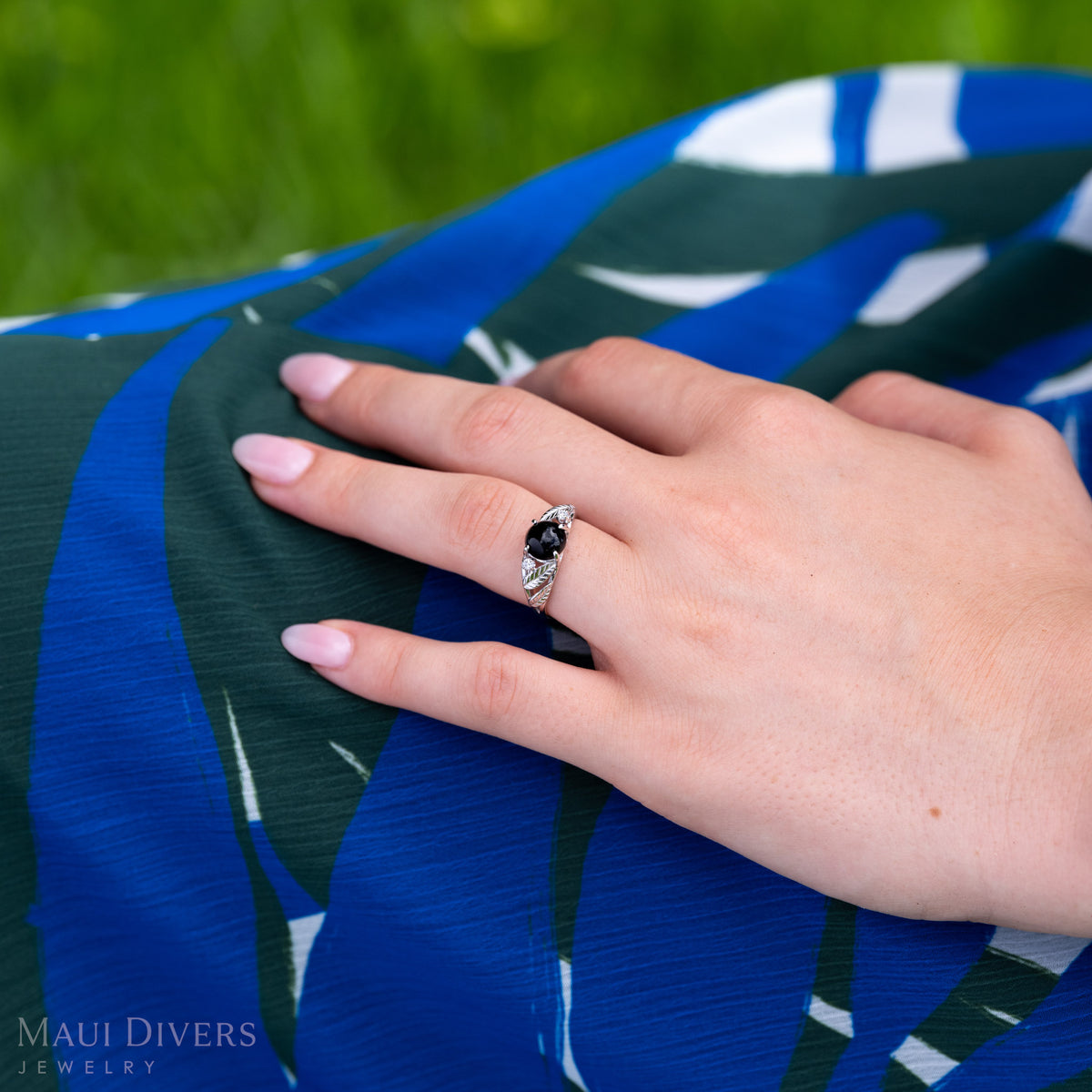 A close-up of a person's hands resting on a blue and green patterned garment, wearing white gold ring with diamond-cut maile leaves, diamond accents, and Hawaiian black coral. The background is a soft-focus green outdoor setting.