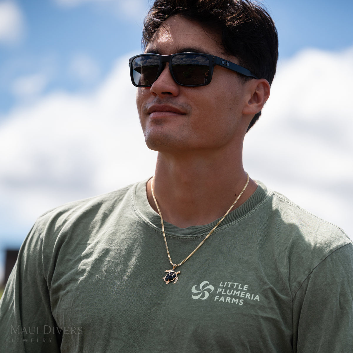 Dane Little wearing a green ʻLittle Plumeria Farmsʻ shirt and gold sea turtle pendant with geometric black coral inlay, against a blue cloudy sky