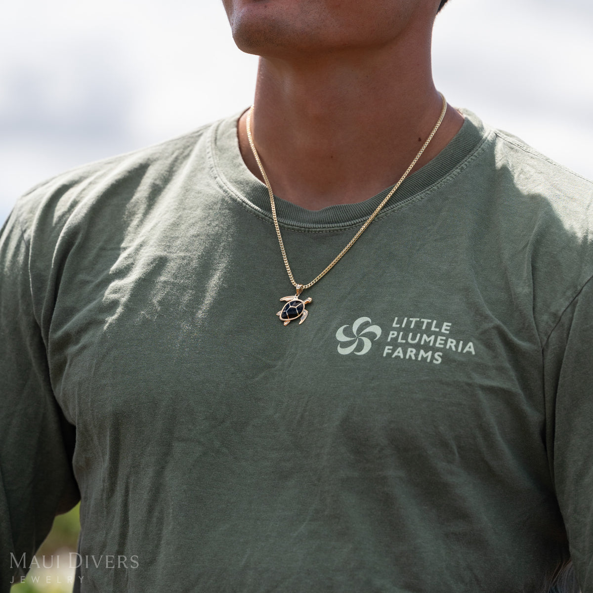 Close up of Dane Little wearing a gold sea turtle necklace with geometric black coral inlay for the shell, green ʻLittle Plumeria Farmsʻ shirt against a sky background