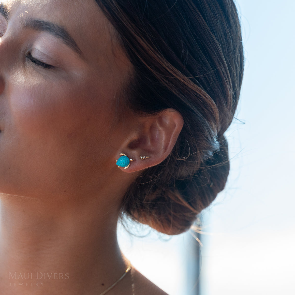 Close-up of a woman's ear with a gold sea turtle ring with turquoise and a small gold conch shape earrings against a sky background