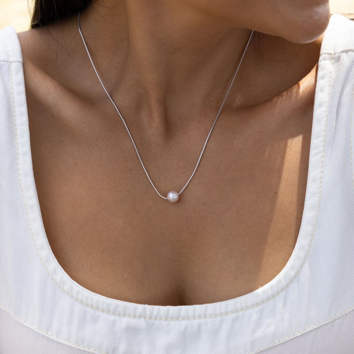 A close-up of a model wearing a delicate silver chain necklace with a single white pearl pendant, styled with a white scoop-neck top.