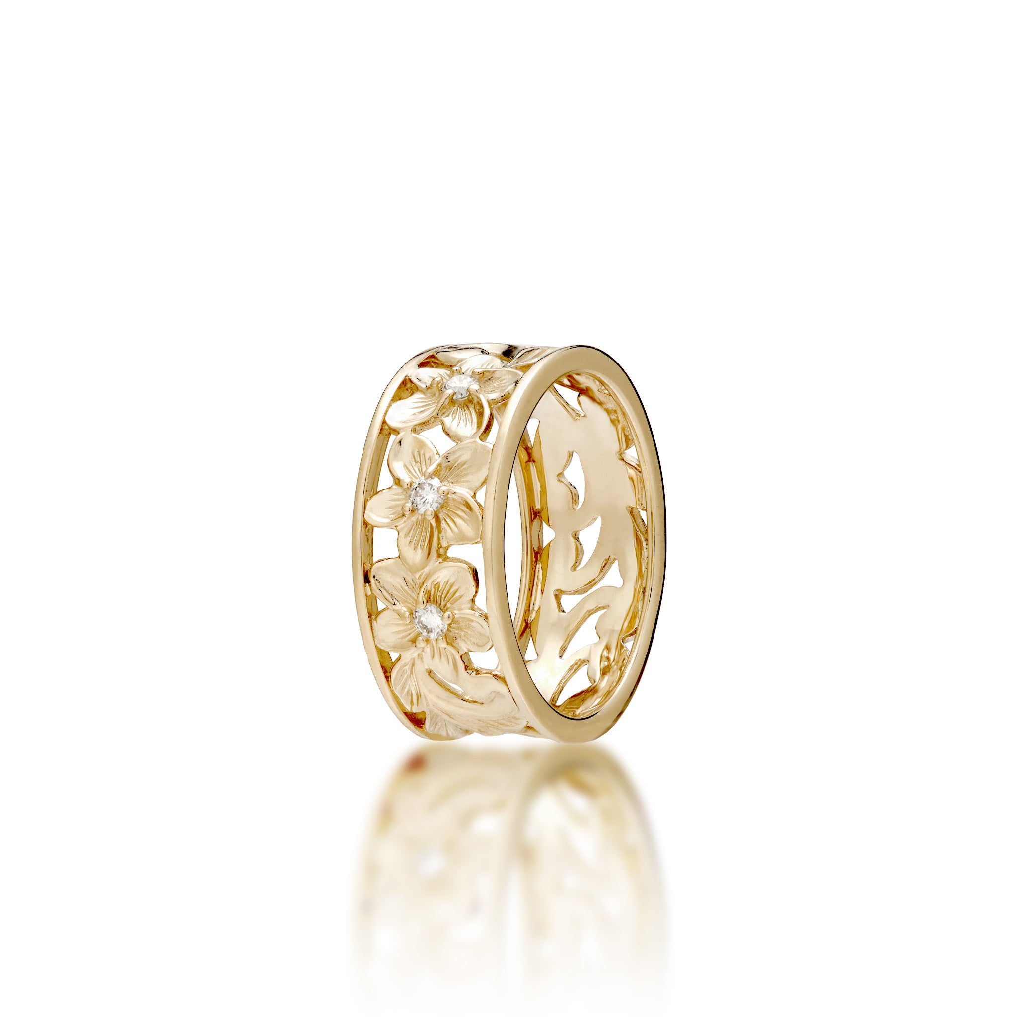 A 14k yellow gold Hawaiian Heirloom band ring featuring an maile leaf and fern scrollwork and plumeria flower pattern with a small diamond at the center of each flower, shown on a white background with a soft reflection below.
