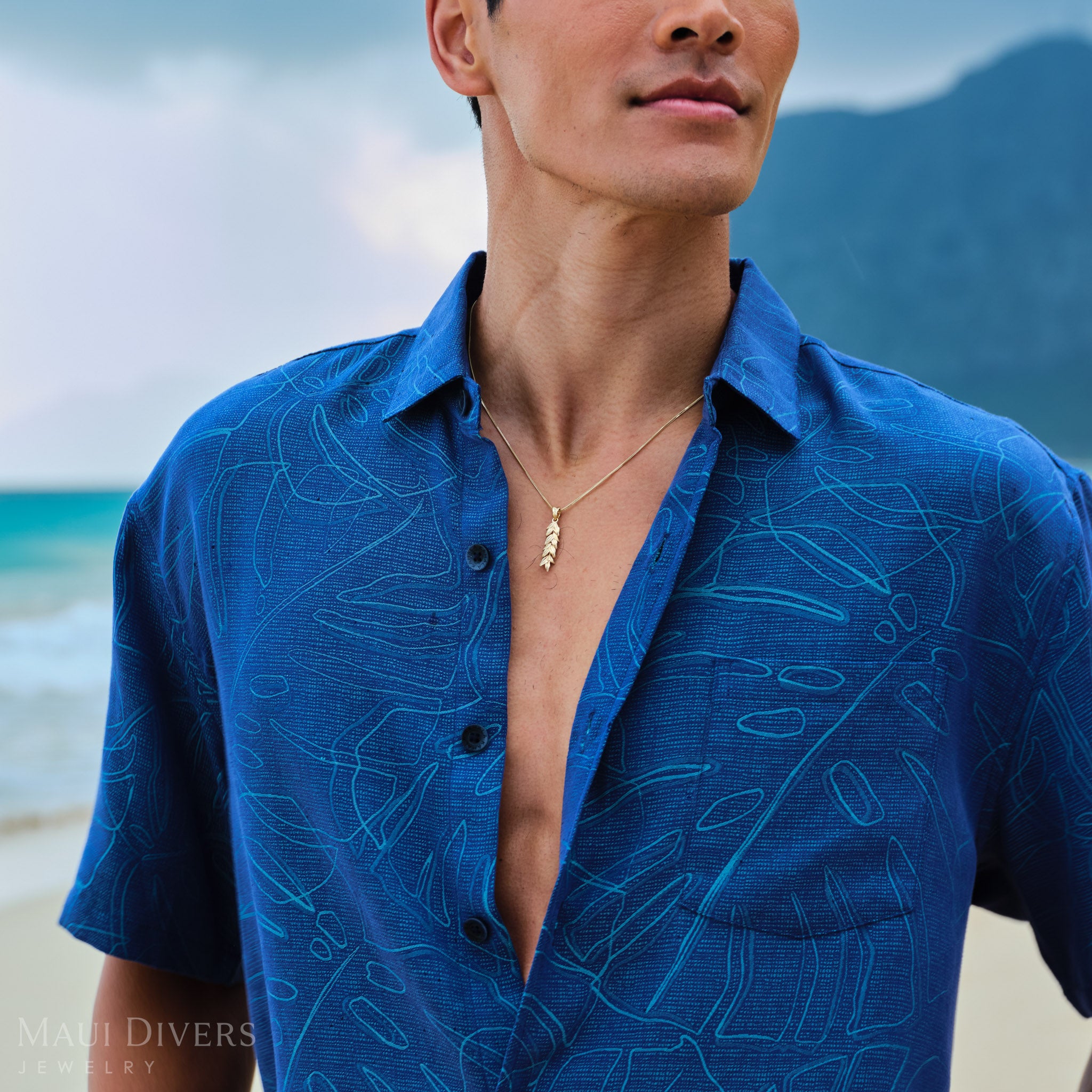 Man wearing a blue patterned shirt  with a gold maile leaf pendant on a chain on a beach with mountains in the background
