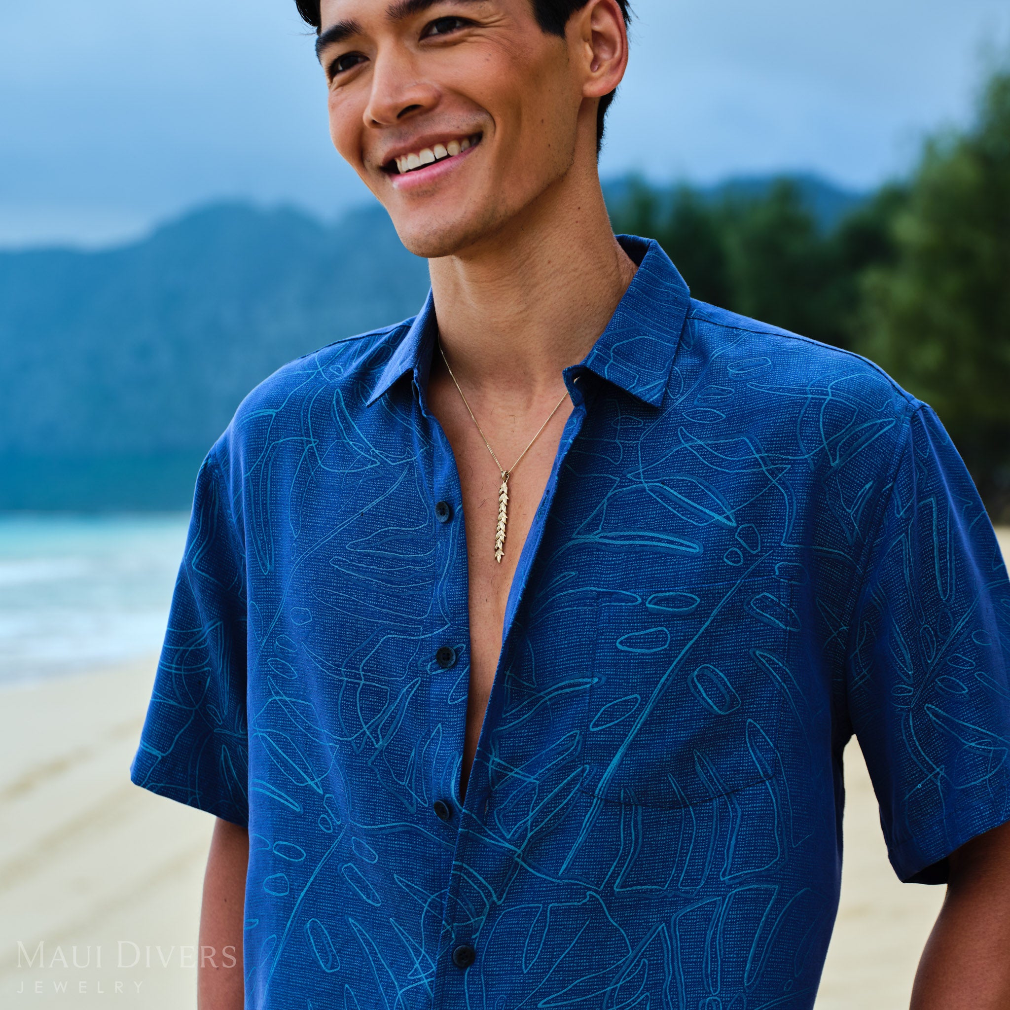Man wearing a gold maile leaf pendant and blue patterned shirt on a beach with mountains in the background