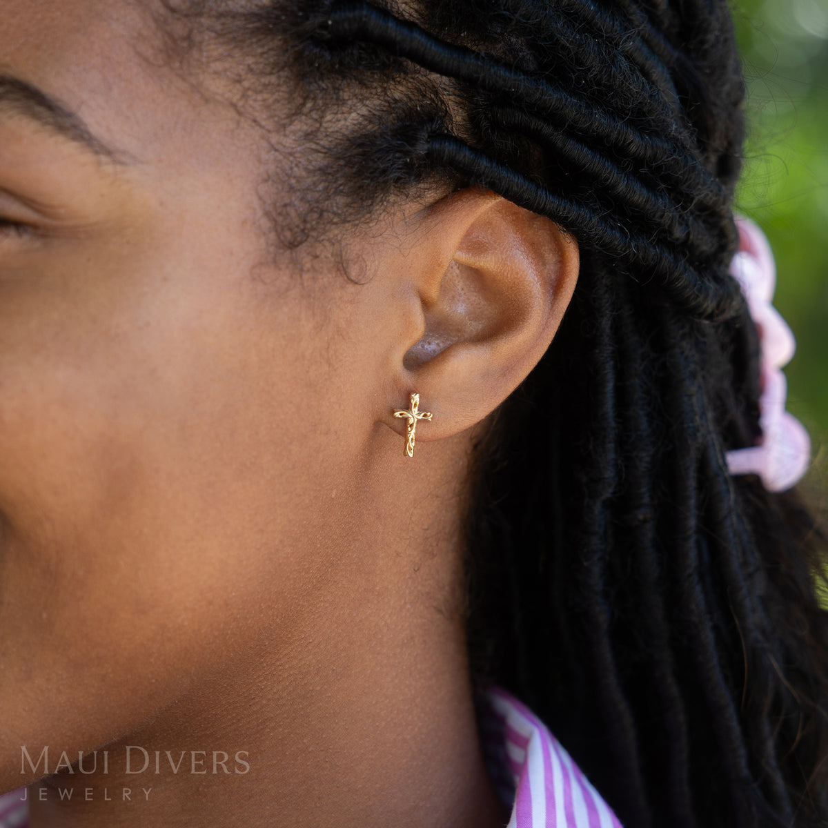 A side view close-up of a woman wearing a Hawaiian Heirloom Old English Scroll Cross Earring in 14k yellow gold, against a blurred leafy green background