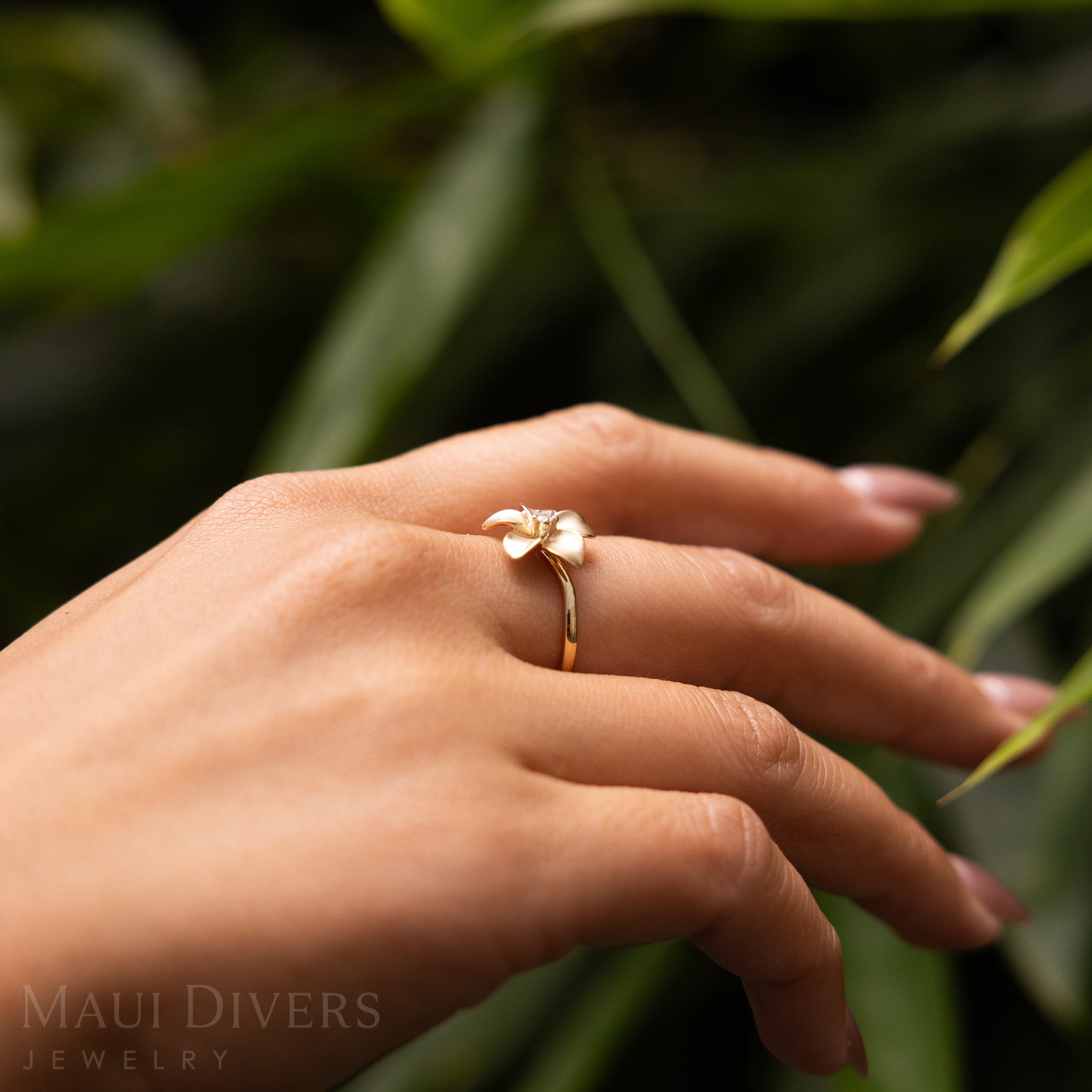 Side view of a hand wearing a Plumeria Ring in 14k yellow gold with a diamond against a blurred leafy background