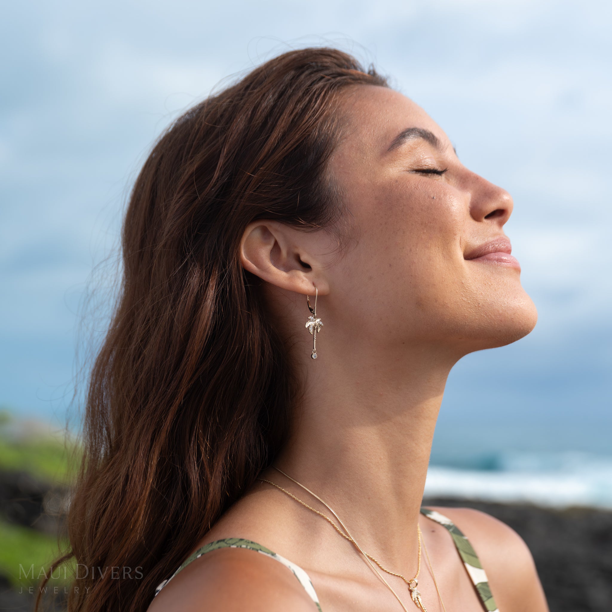 Woman wearing gold and diamond palm tree earrings with a blue sky in the background