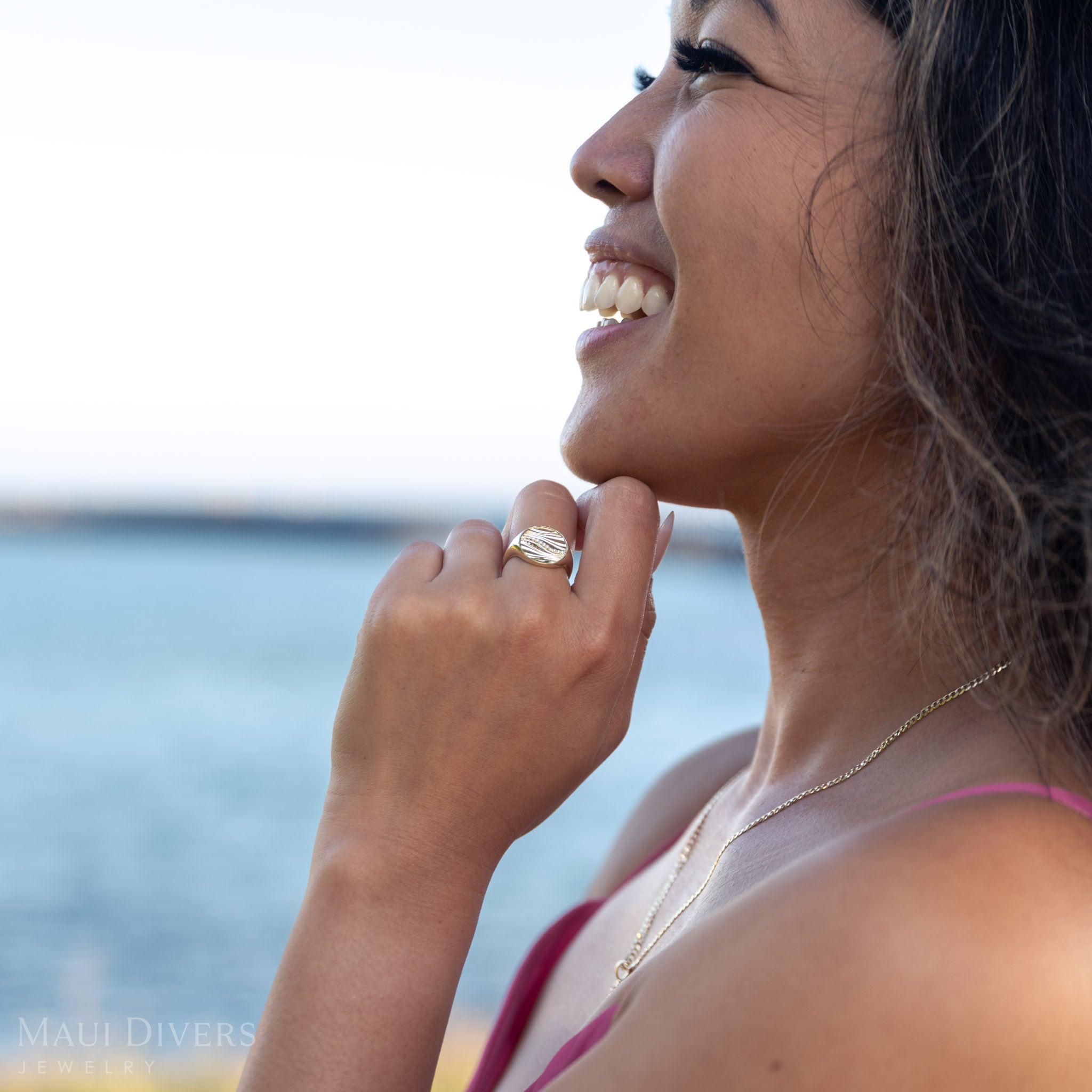 Woman smiling wearing a ocean sand ring in 14k yellow gold with a blurred background of water and sky