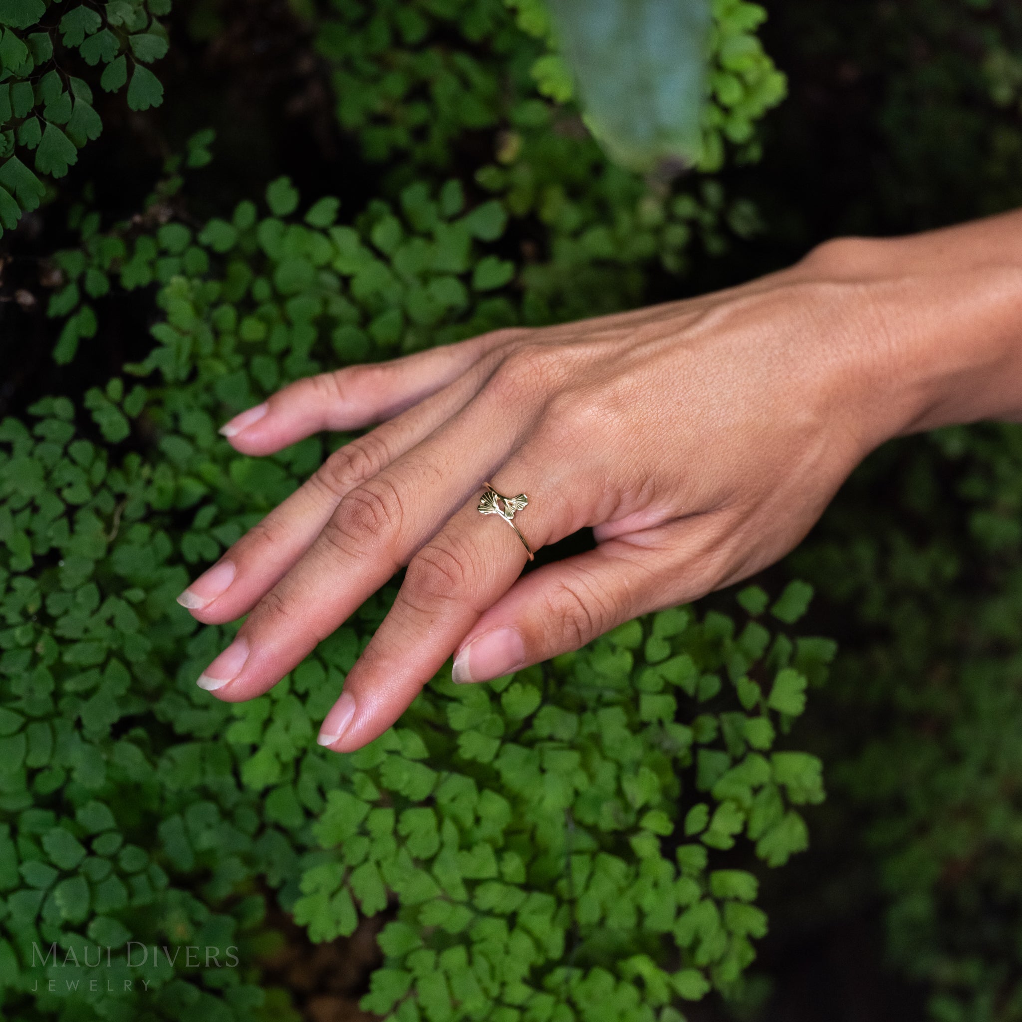 A close-up of a person's hand wearing a delicate maiden hair fern ring in 14k yellow gold, set against a background of lush green fern leaves.