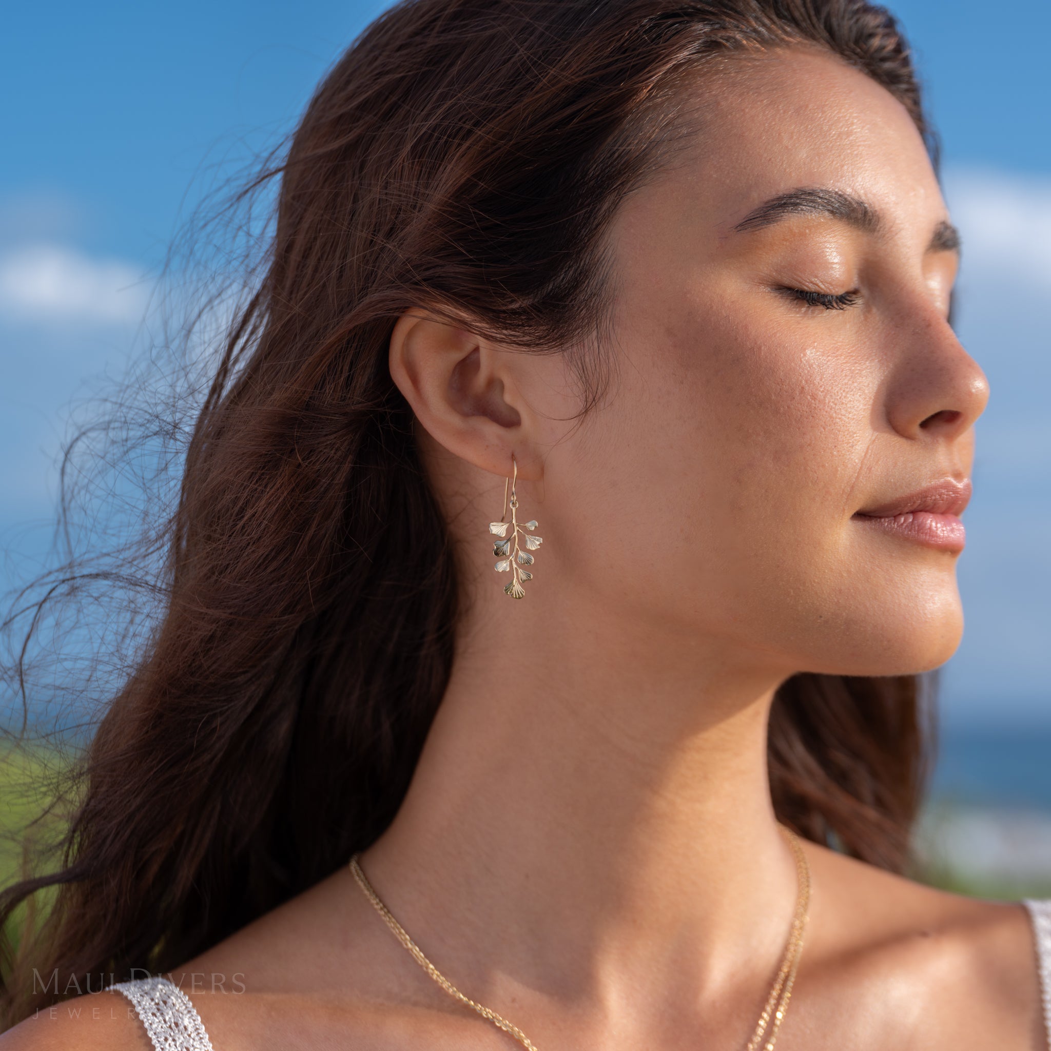 A woman wearing a delicate Maiden Hair 14k yellow gold dangle hook earring, set against a blurred outdoor background of blue sky and greenery.