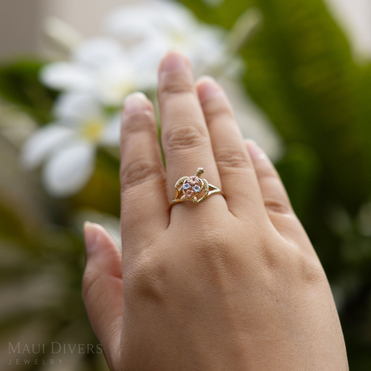 A 14k gold Hawaiian green sea turtle ring with two rose and two white gold plumerias on its back, shown on a hand with blurry plumeria flowers in the background