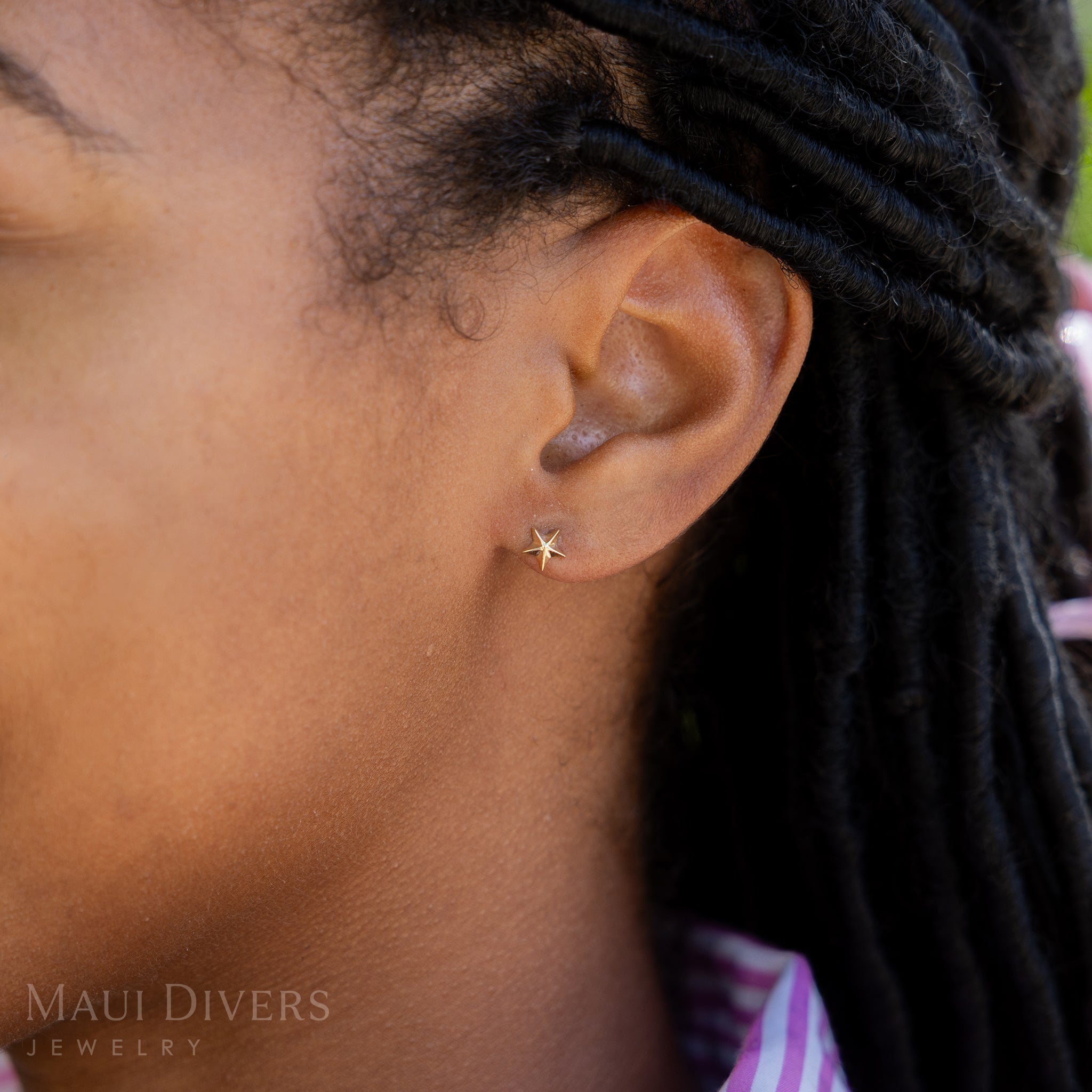 A side view close-up of a woman wearing a Starfish Earring in 14k yellow gold, against a blurred leafy green background