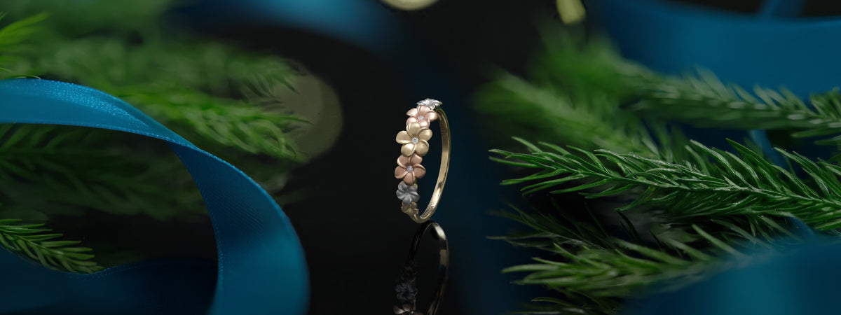A delicate plumeria flower ring with yellow, rose, and white gold plumeria with diamonds, standing upright against a dark background with green pine branches and a blue ribbon.