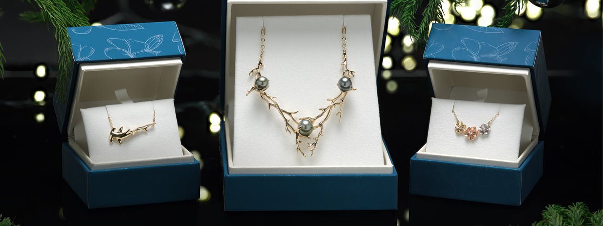 Three jewelry gift boxes are arranged on a dark surface, with the center box open to display a gold coral branch-style necklace with Tahitian pearls, and the side boxes open to display a tri color gold plumeria flower necklace and shark necklace