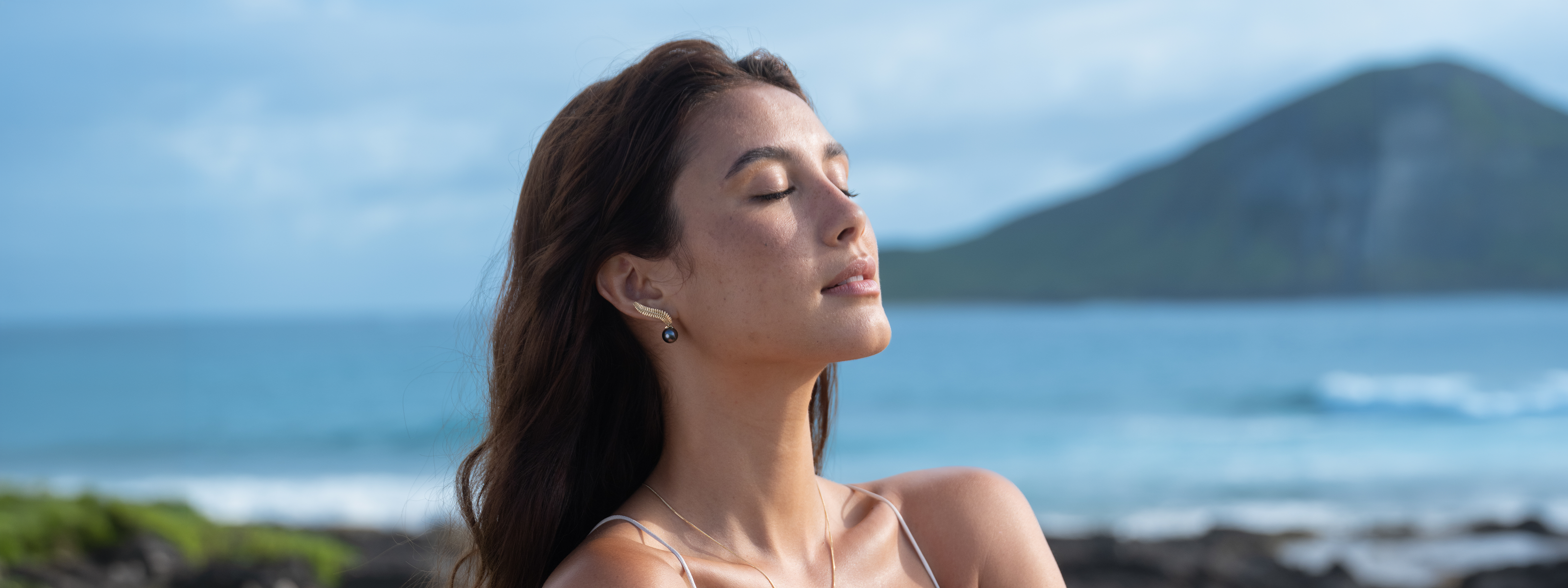 Woman with eyes closed standing  and wearing a kupukupu fern Tahitian black pearl earring on a beach with mountains in the background