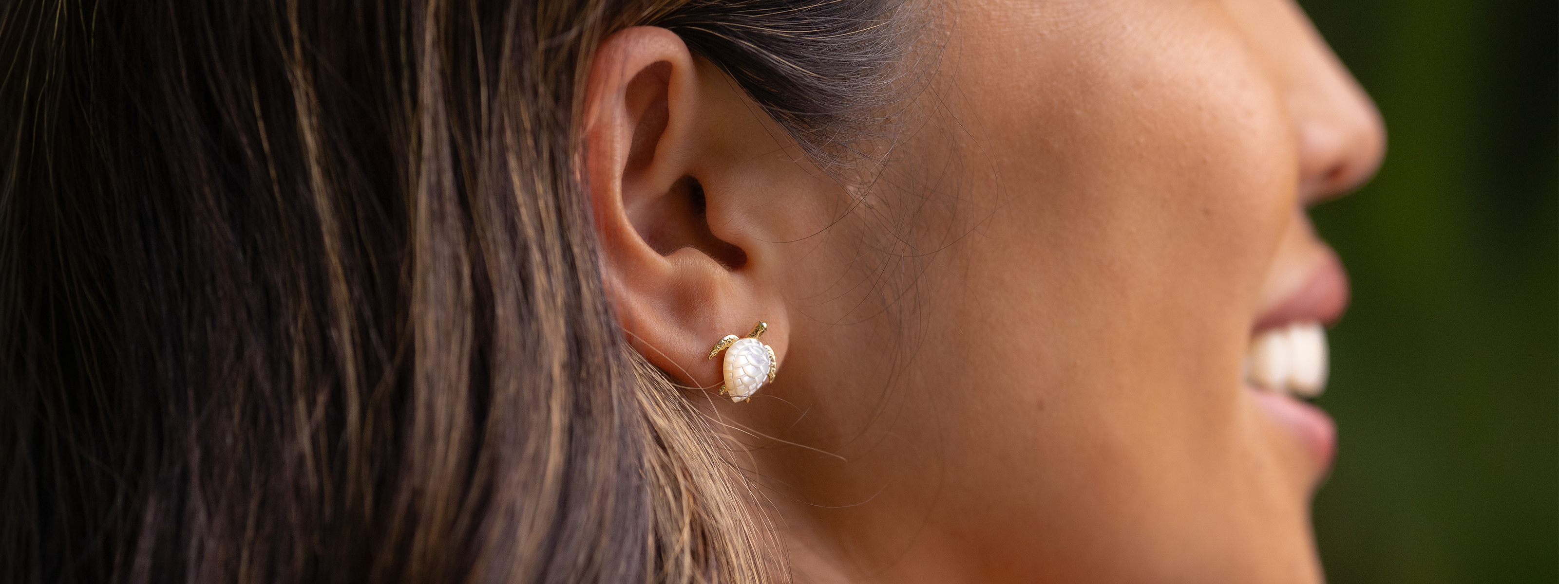 A close-up side profile of a woman wearing Hawaiian Green Sea Turtle Earrings with a white mother-of-pearl inlay for the shell design, set against a blurred green background.