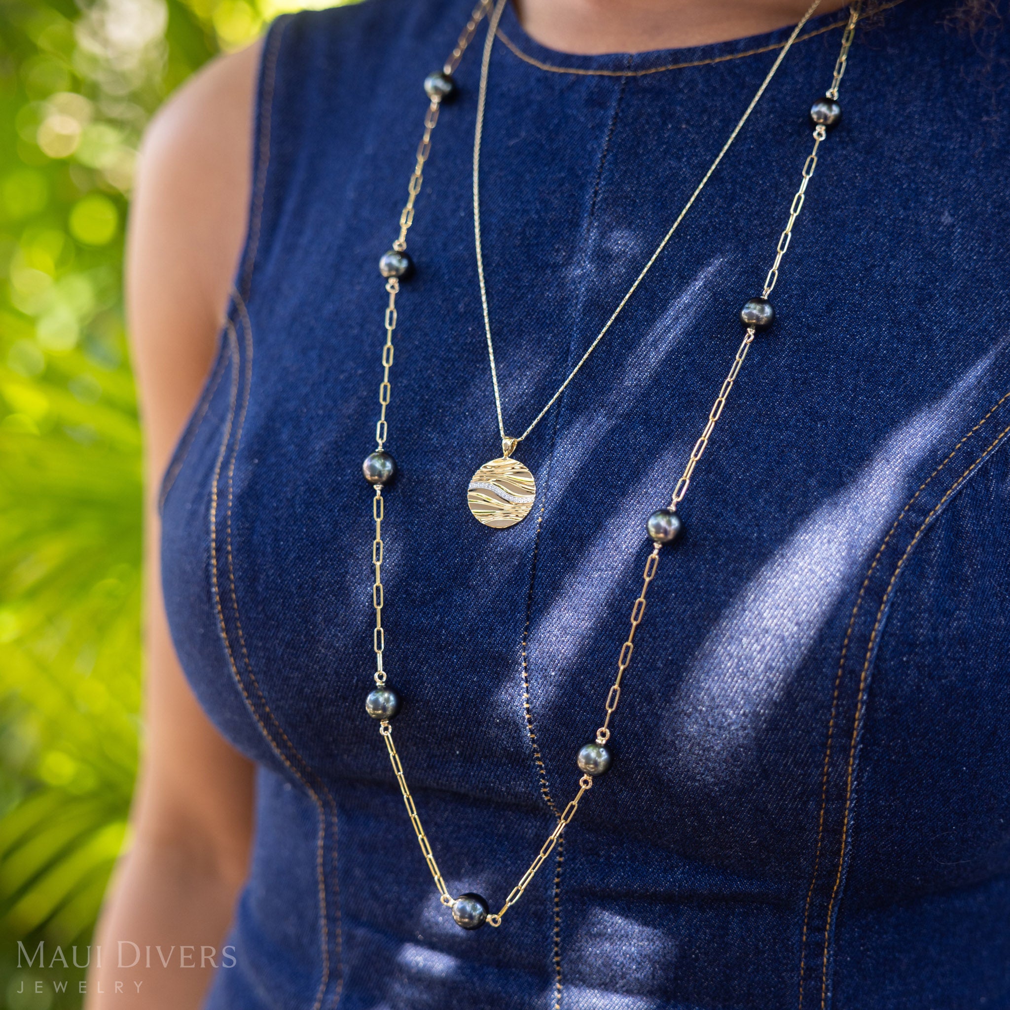 A person wearing a long Tahitian black pearl necklace and the Ocean Sand Pendant with a blue top outdoors