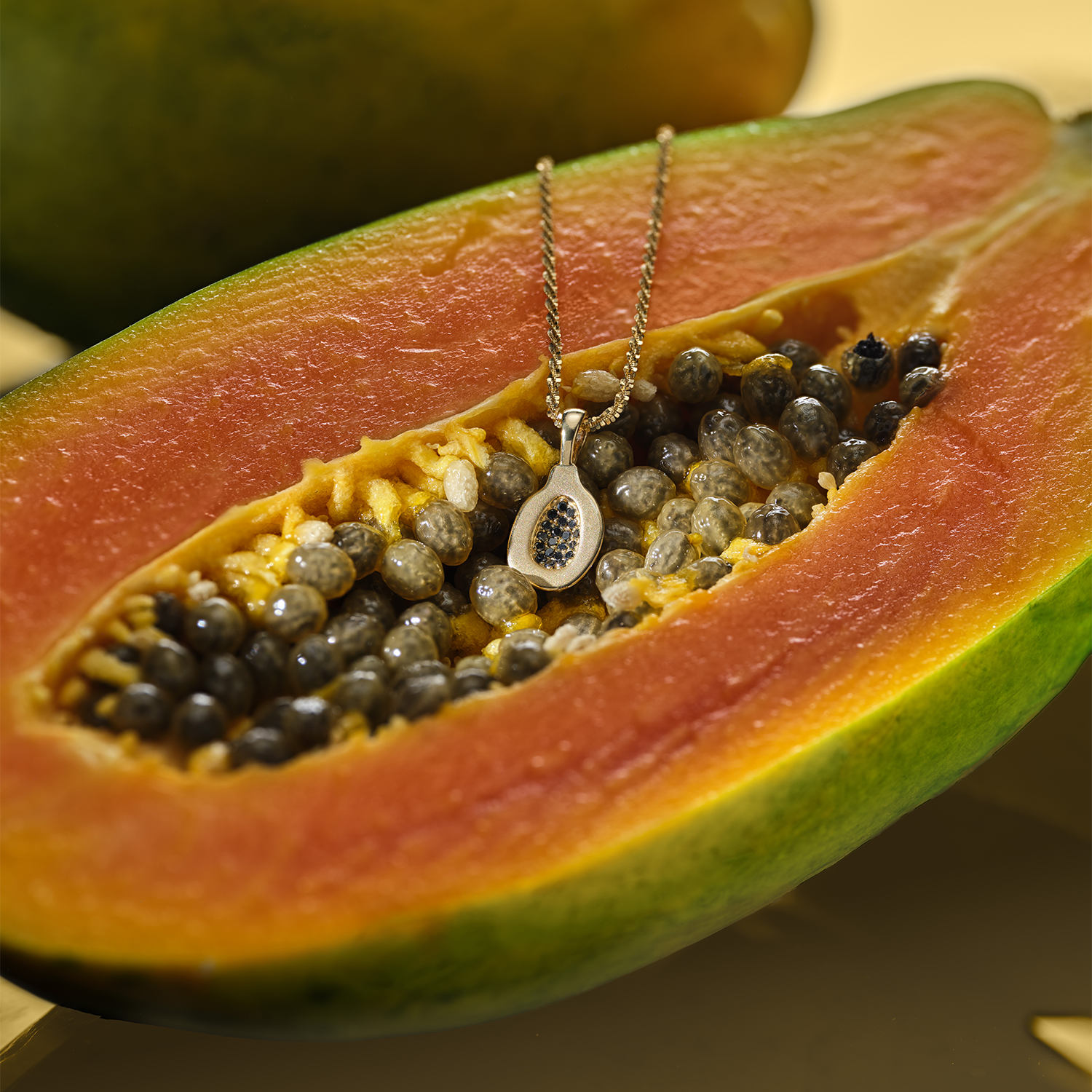 Gold Papaya Pendant with black diamonds on a chain resting atop a half-cut papaya with a blurry papaya in the background