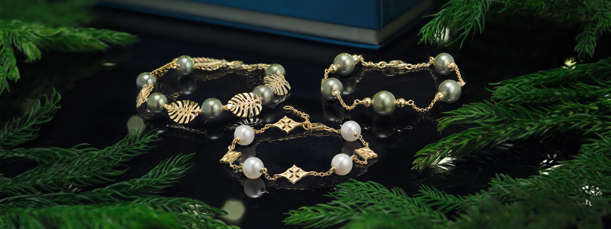 Three pearl bracelets in 14k yellow gold arranged on a glossy black surface surrounded by green pine branches. The back two feature Tahitian black pearls, and the middle features akoya white pearls