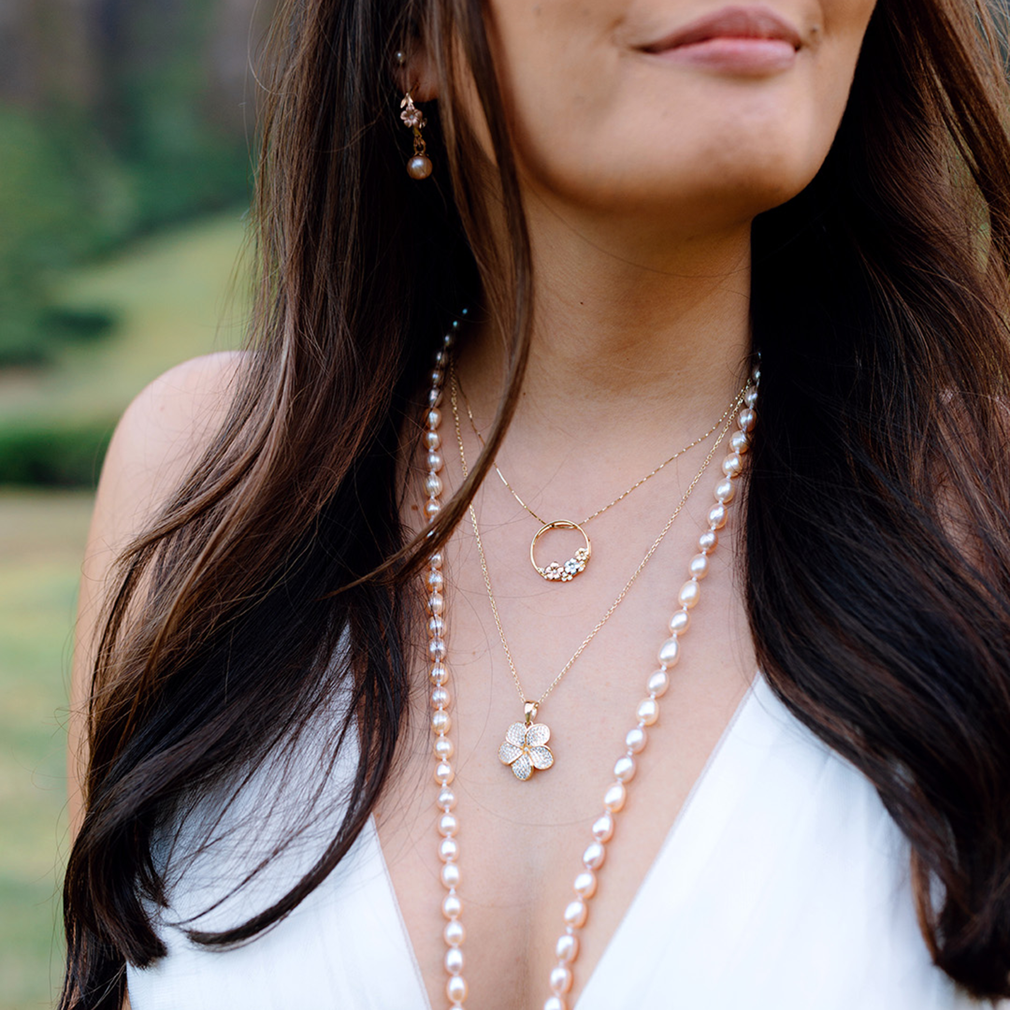 Close-up of a woman wearing a pearl necklace, two plumeria necklaces and earrings with a blurred natural background