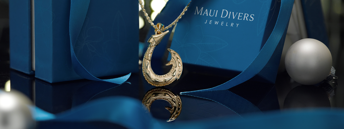 A gold Maui fish hook pendant on a rope chain displayed in front of a blue Maui Divers Jewelry box and ribbon, with reflective dark surfaces and a white decorative ornament nearby.
